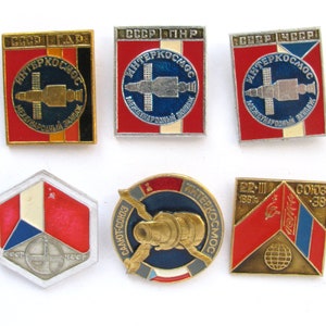 May include: Six enamel pins featuring the Interkosmos logo, a space station with the text "Interkosmos" and "International Crew" in Russian. The pins are in various colors, including red, blue, white, and gold. The pins are arranged in two rows of three.