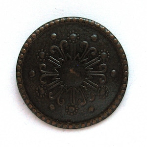 May include: A round, antique-style button with a dark bronze finish. The button features an ornate, symmetrical design in the center, surrounded by a dotted border. The design includes floral and geometric elements.