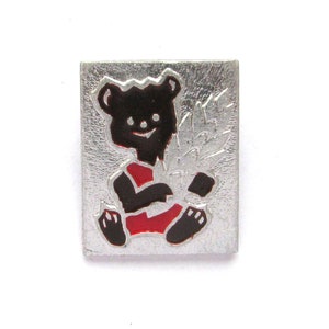 Op de afbeelding: Een zilveren pin met een zwarte beer die een rood shirt draagt en een wit blad vasthoudt. De beer glimlacht en heeft een vriendelijke uitdrukking.