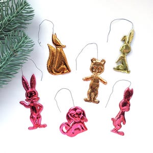 Op de afbeelding: Een verzameling vintage kerstversieringen in de vorm van dieren. De ornamenten omvatten een gouden eekhoorn, een gouden beer, een gouden konijn, en drie roze konijnen en een roze hond. Elk ornament heeft een draadlus om op te hangen.