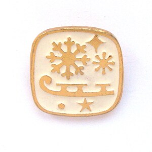 May include: A square pin with a gold border and a white interior. The pin features a gold snowflake, two stars, and a sled design. The pin is likely a winter or holiday-themed accessory.