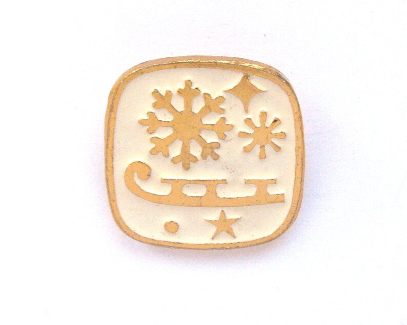 May include: A square pin with a gold border and a white interior. The pin features a gold snowflake, two stars, and a sled design. The pin is likely a winter or holiday-themed accessory.