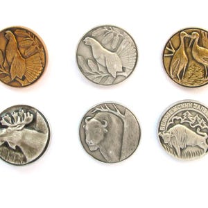 May include: Six round metal buttons with embossed wildlife designs. The buttons feature images of a bird, moose, bison, and other animals. The buttons are in shades of gold and silver, with intricate details.