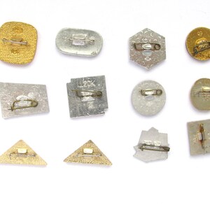 May include: A collection of vintage metal pins in various shapes, including squares, circles, and triangles. The pins have a gold or silver finish and feature pin backs for attachment. Some pins have text or designs.