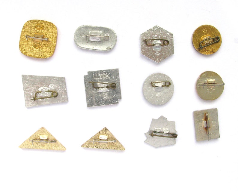 May include: A collection of vintage metal pins in various shapes, including squares, circles, and triangles. The pins have a gold or silver finish and feature pin backs for attachment. Some pins have text or designs.