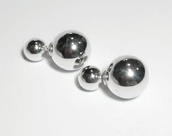 May include: A pair of shiny silver double-sided ball earrings. Each earring features a small ball connected to a larger ball. The earrings are displayed on a white surface, reflecting light.