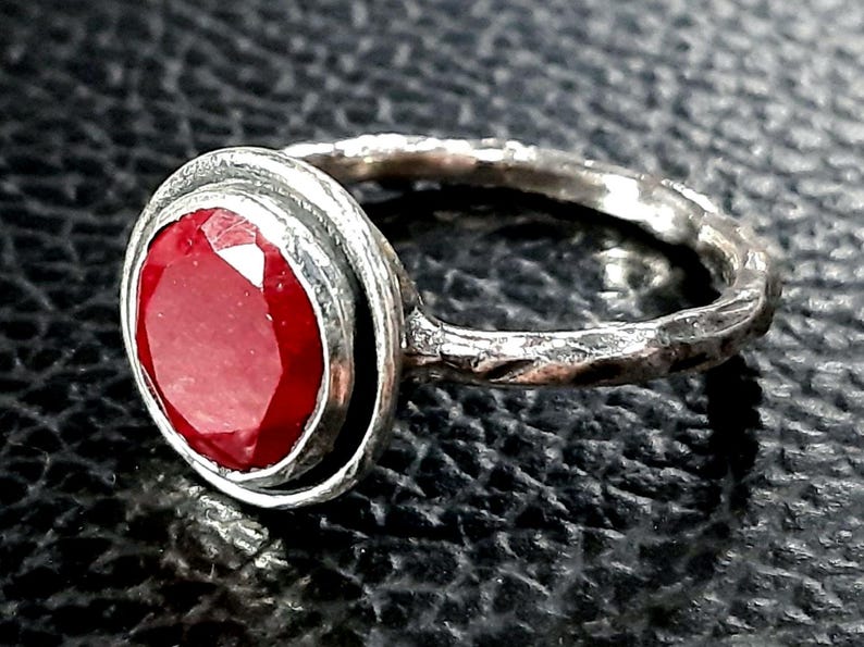 Pu&ograve; includere: Un anello d'argento con una grande pietra preziosa rossa rotonda incastonata in una montatura a castone. La fascia dell'anello &egrave; testurizzata e ha una finitura martellata.