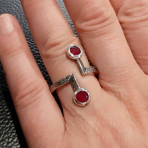 Op de afbeelding: Een zilveren ring met twee rode edelstenen. De ring heeft een geometrisch ontwerp met een getextureerde band.