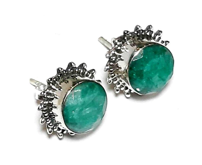 Puede incluir: Un par de pendientes de bot&oacute;n con piedras preciosas redondas, facetadas, de color verde esmeralda. Las piedras preciosas est&aacute;n engastadas en un metal plateado con un borde decorativo y puntiagudo. Sobre un fondo blanco.