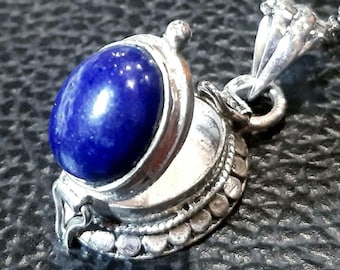 May include: A silver pendant necklace featuring a deep blue, oval-shaped gemstone. The gemstone is set in a detailed, ornate silver setting. The necklace chain is also silver and appears delicate. The pendant has a vintage or antique aesthetic.
