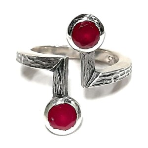 Op de afbeelding: Een zilveren ring met een getextureerde band en twee rode edelstenen. De ring is aan &eacute;&eacute;n kant open en heeft een uniek ontwerp.