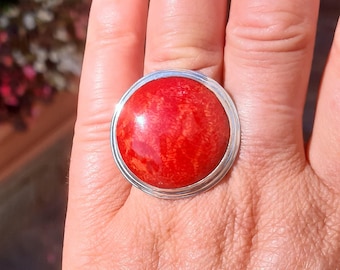 May include: A ring featuring a large, round, polished red stone set in a smooth, silver-toned metal bezel. The stone displays a vibrant, saturated red hue with subtle, natural-looking organic textures and patterns visible across its surface.