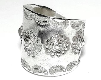 May include: A wide, silver-toned ring with intricate floral and leaf patterns. The ring features embossed designs of spiraling flowers and detailed leaves, creating a textured surface. The ring's design is bold and eye-catching, suitable for a statement piece.