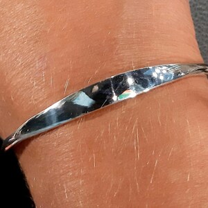 May include: A silver bangle bracelet with a smooth, curved design. The bracelet is worn on a person's wrist.