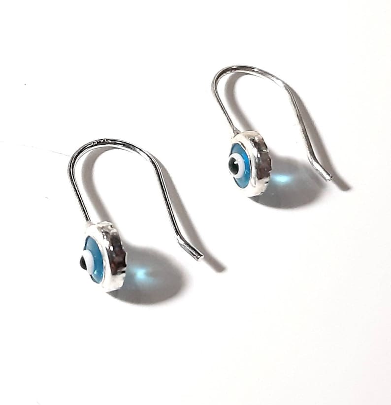 K&ouml;nnte beinhalten: Ein Paar silberne Ohrringe mit blauen b&ouml;sen Augen-Anh&auml;ngern. Die Anh&auml;nger sind rund und haben eine wei&szlig;e Mitte mit einer schwarzen Pupille. Die Ohrringe befinden sich auf einem wei&szlig;en Hintergrund.
