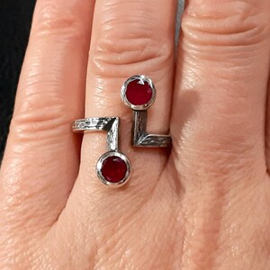 Op de afbeelding: Een zilveren ring met twee rode edelstenen die in de band zijn gezet. De ring is ontworpen met een geometrische vorm en heeft een getextureerde afwerking.