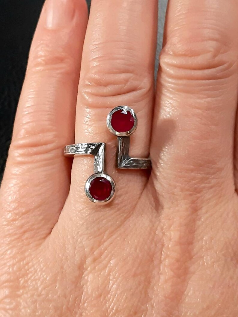 Op de afbeelding: Een zilveren ring met twee rode edelstenen die in de band zijn gezet. De ring is ontworpen met een geometrische vorm en heeft een getextureerde afwerking.