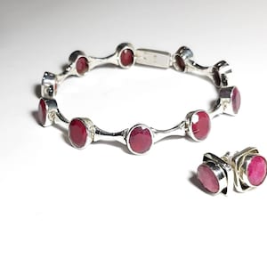 May include: A silver bracelet with red gemstones and matching earrings. The bracelet has a hinged clasp and is decorated with small silver accents. The earrings are stud style with square settings.