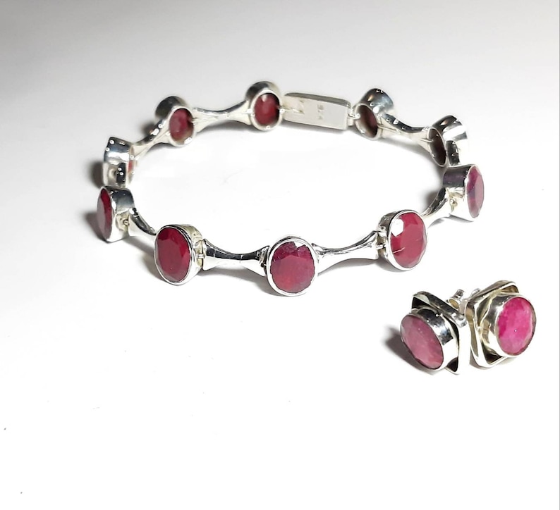 May include: A silver bracelet with red gemstones and matching earrings. The bracelet has a hinged clasp and is decorated with small silver accents. The earrings are stud style with square settings.
