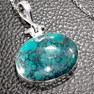Puede incluir: Un collar con un colgante de plata y una piedra turquesa. La piedra es de color azul claro con vetas azul oscuro que la atraviesan. El colgante tiene forma ovalada y un acabado liso y pulido.