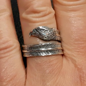 May include: A silver-colored ring featuring an eagle head design. The ring wraps around the finger with a feather-like band. The eagle's head is detailed with textured feathers and a sharp beak.