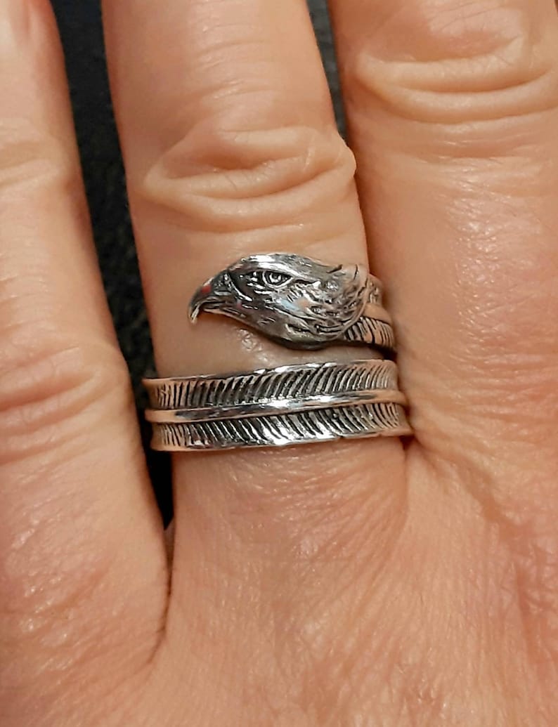 May include: A silver-colored ring featuring an eagle head design. The ring wraps around the finger with a feather-like band. The eagle's head is detailed with textured feathers and a sharp beak.