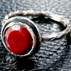 Pu&ograve; includere: Un anello d'argento con una pietra preziosa rossa incastonata in una montatura a castone. La fascia dell'anello &egrave; testurizzata e ha una finitura martellata.