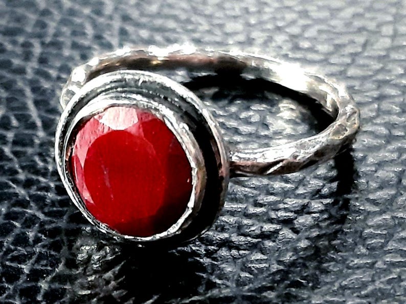Pu&ograve; includere: Un anello d'argento con una pietra preziosa rossa incastonata in una montatura a castone. La fascia dell'anello &egrave; testurizzata e ha una finitura martellata.