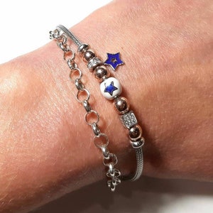 Puede incluir: Un brazalete de cadena de plata con un brazalete de cadena de oro rosa superpuesto. El brazalete de oro rosa tiene una cuenta blanca con una estrella azul en ella, una cuenta de oro rosa y una cuenta de forma cuadrada con piedras transparentes. Un peque&ntilde;o amuleto de estrella azul cuelga de la cadena de oro rosa.