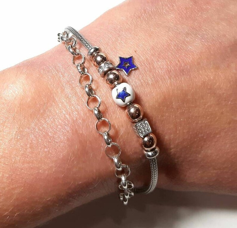 Puede incluir: Un brazalete de cadena de plata con un brazalete de cadena de oro rosa superpuesto. El brazalete de oro rosa tiene una cuenta blanca con una estrella azul en ella, una cuenta de oro rosa y una cuenta de forma cuadrada con piedras transparentes. Un peque&ntilde;o amuleto de estrella azul cuelga de la cadena de oro rosa.