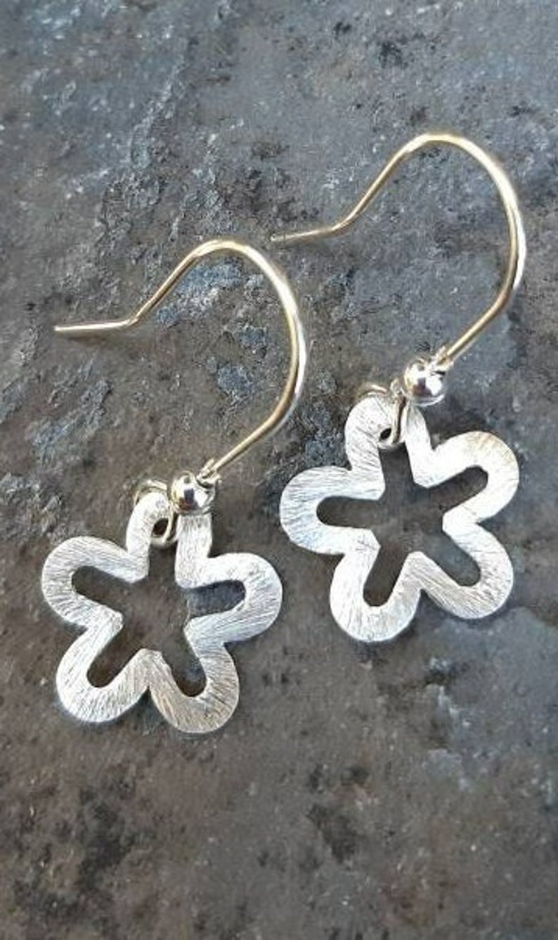 K&ouml;nnte beinhalten: Ein Paar silberfarbene, bl&uuml;tenf&ouml;rmige Ohrringe mit geb&uuml;rsteter Metalloberfl&auml;che. Die Ohrringe haben ein Haken-Design und werden auf einer strukturierten, dunkelgrauen Oberfl&auml;che pr&auml;sentiert. Das florale Design hat f&uuml;nf Bl&uuml;tenbl&auml;tter.