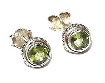 May include: A pair of stud earrings featuring round, faceted, light green peridot gemstones set in polished silver. Each earring has a textured, rope-style border surrounding the stone and includes a standard silver butterfly-style earring back.
