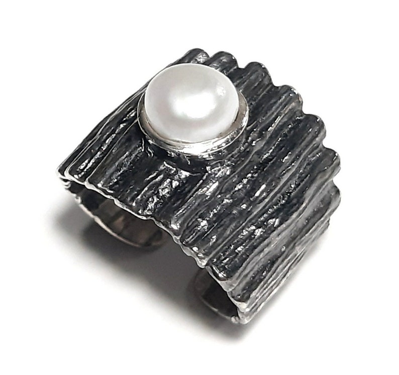 May include: A silver ring with a textured, wood-like band and a white pearl set in the center.