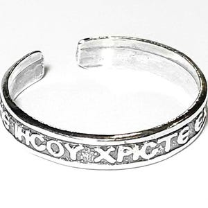 May include: A silver-colored cuff bracelet with an open design. The bracelet features engraved text around the band. The text appears to be in a foreign language. The bracelet is set against a white background.