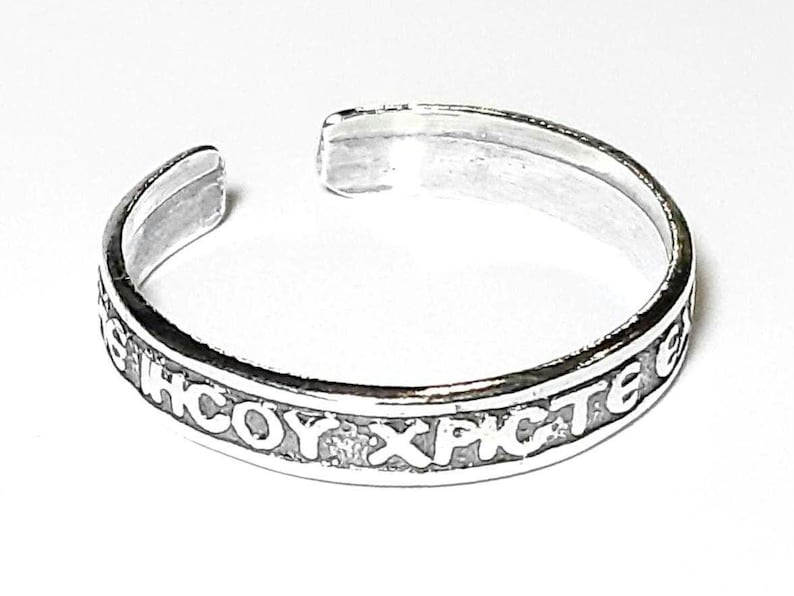 May include: A silver-colored cuff bracelet with an open design. The bracelet features engraved text around the band. The text appears to be in a foreign language. The bracelet is set against a white background.