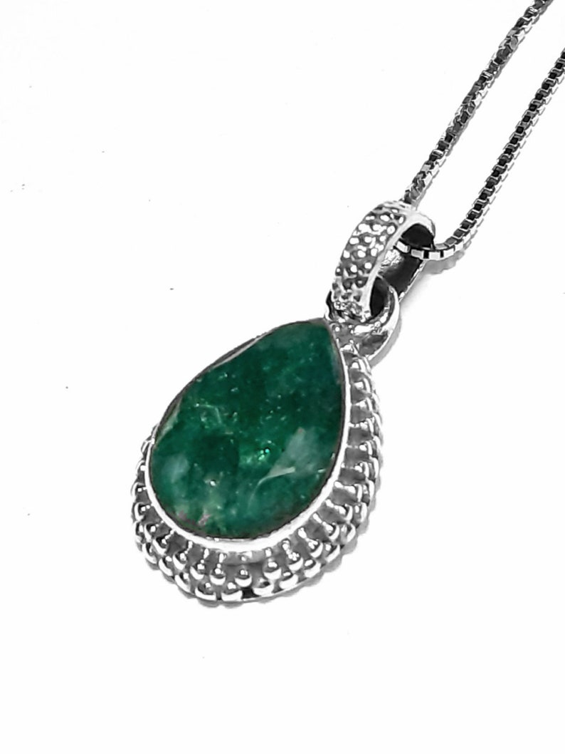 May include: A silver necklace with a teardrop-shaped pendant featuring a green gemstone set in a silver bezel. The pendant is surrounded by a decorative silver border.