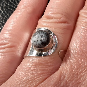 May include: A silver ring with a black and white stone set in a bezel setting. The ring is on a person's finger.