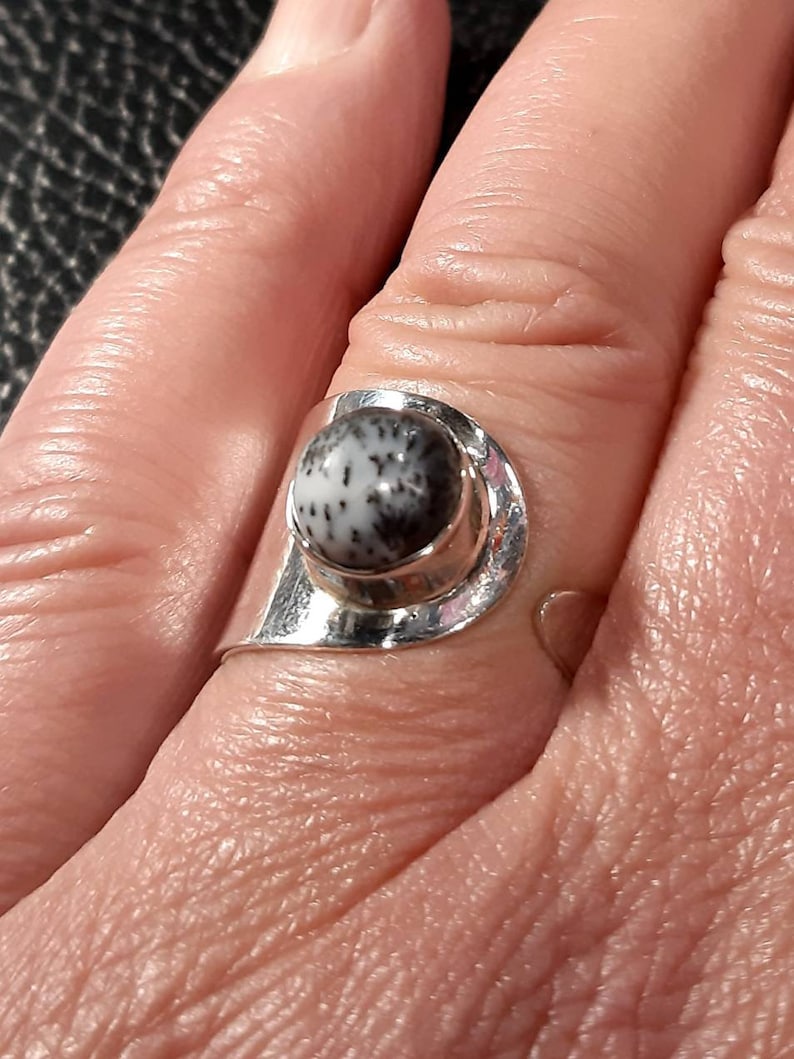 May include: A silver ring with a black and white stone set in a bezel setting. The ring is on a person's finger.