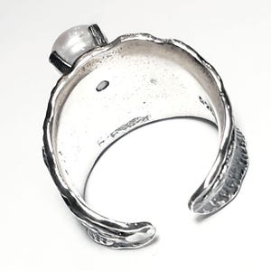 May include: A silver ring with a textured band and a white pearl set in the top. The ring is open on one side and has a slightly curved shape.