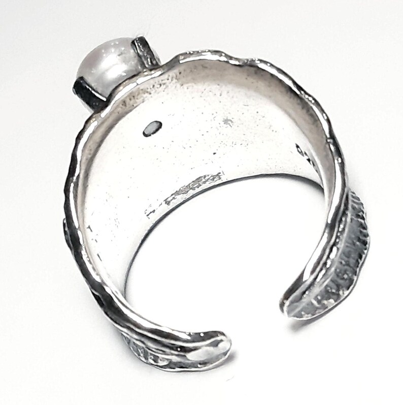 May include: A silver ring with a textured band and a white pearl set in the top. The ring is open on one side and has a slightly curved shape.