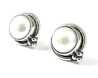 May include: A pair of stud earrings featuring large, round, white pearls set in a silver metal. The earrings have a decorative, swirling design around the pearl and small, round accents at the base.