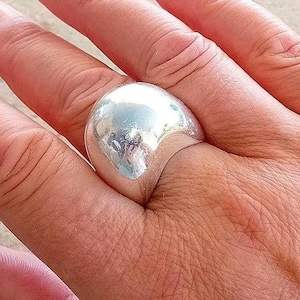 Puede incluir: Un anillo de tono plateado pulido que presenta una superficie lisa, redondeada y abovedada. El metal tiene un acabado altamente reflectante que capta la luz, creando un brillo metálico intenso sobre la forma curva y bulbosa del aro.