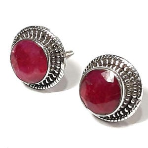Peut inclure: Une paire de boucles d'oreilles à clous avec de grosses pierres précieuses rondes, facettées et rouge foncé. Les pierres précieuses sont serties dans un sertissage à lunette argenté et texturé avec une bordure décorative.