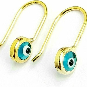 K&ouml;nnte beinhalten: Ein Paar goldene Ohrringe mit blauen b&ouml;sen Augen-Anh&auml;ngern. Die Anh&auml;nger sind rund und haben eine wei&szlig;e Mitte mit einer schwarzen Pupille.