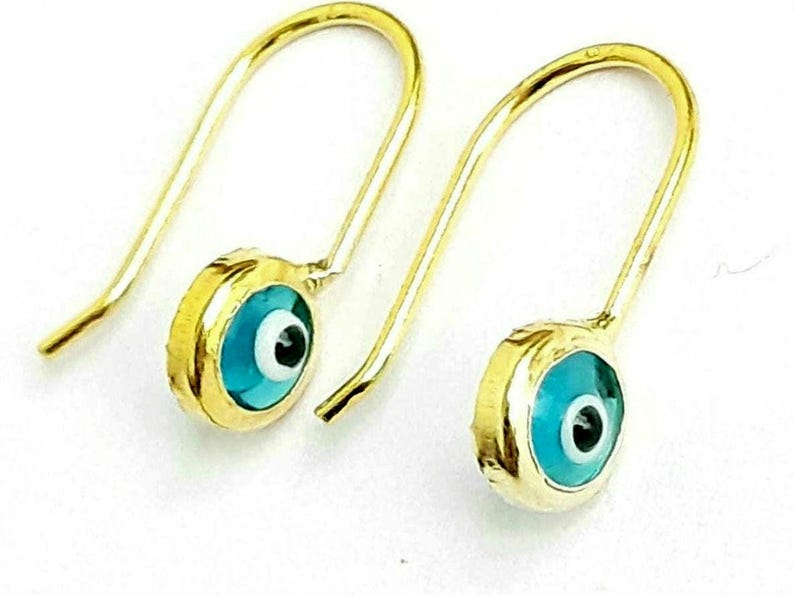 K&ouml;nnte beinhalten: Ein Paar goldene Ohrringe mit blauen b&ouml;sen Augen-Anh&auml;ngern. Die Anh&auml;nger sind rund und haben eine wei&szlig;e Mitte mit einer schwarzen Pupille.