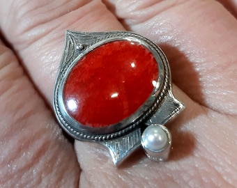 May include: A silver ring featuring a large, oval, bright red stone. The stone is surrounded by a detailed silver setting with a leaf-like design. A small, round pearl is set on the side of the ring.