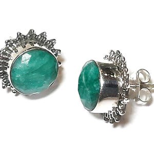 Puede incluir: Un par de pendientes de bot&oacute;n con piedras preciosas verde esmeralda ovaladas y facetadas engastadas en metal plateado. El engaste met&aacute;lico tiene un dise&ntilde;o texturizado en forma de sol alrededor de la piedra. Sobre un fondo blanco.