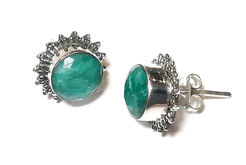 Puede incluir: Un par de pendientes de bot&oacute;n con piedras preciosas verde esmeralda ovaladas y facetadas engastadas en metal plateado. El engaste met&aacute;lico tiene un dise&ntilde;o texturizado en forma de sol alrededor de la piedra. Sobre un fondo blanco.