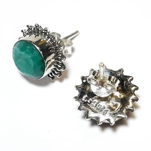 Puede incluir: Dos pendientes de plata con cierre de tuerca. Cada pendiente presenta una piedra preciosa verde esmeralda redonda y facetada engastada en un engaste de plata decorativo. Los engastes tienen un dise&ntilde;o texturizado en forma de sol. Un pendiente se muestra desde el frente, el otro desde la parte posterior.