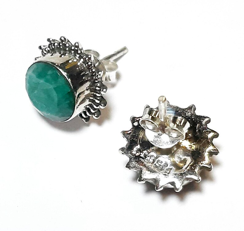 Puede incluir: Dos pendientes de plata con cierre de tuerca. Cada pendiente presenta una piedra preciosa verde esmeralda redonda y facetada engastada en un engaste de plata decorativo. Los engastes tienen un dise&ntilde;o texturizado en forma de sol. Un pendiente se muestra desde el frente, el otro desde la parte posterior.